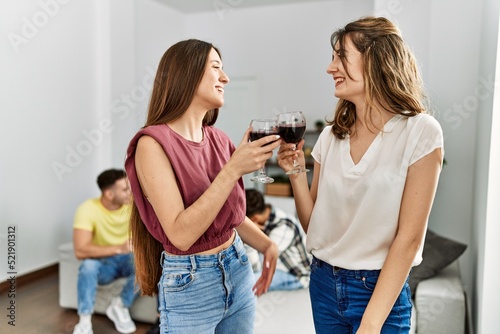 Group of young friends smiling happy sitting on the sofa. Two women toasting with glass of red wine at home.