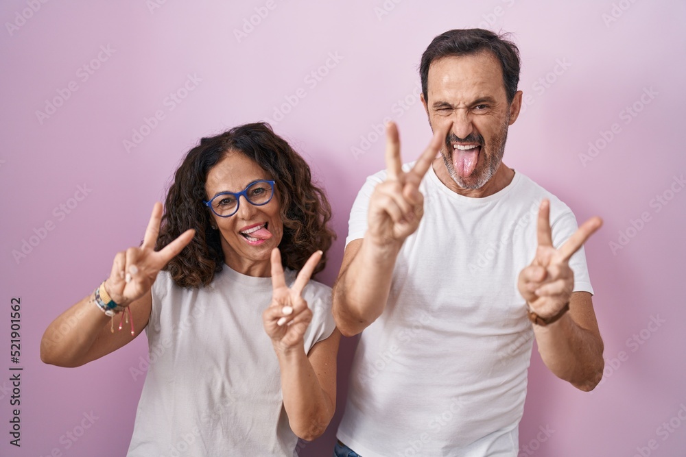 Middle age hispanic couple together over pink background smiling with tongue out showing fingers of both hands doing victory sign. number two. Middle age hispanic couple together over pink background smiling with tongue out showing fingers of both hands doing victory sign. number two.