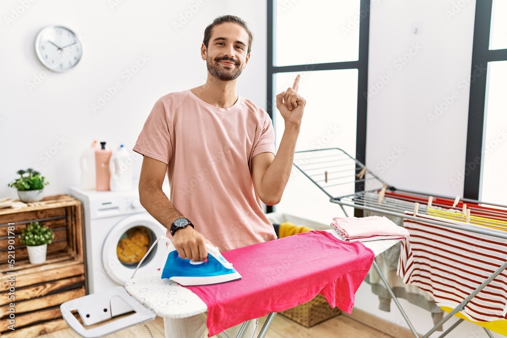 Obraz premium Young hispanic man ironing clothes at home with a big smile on face, pointing with hand finger to the side looking at the camera.