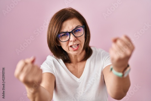Middle age hispanic woman standing over pink background angry and mad raising fists frustrated and furious while shouting with anger. rage and aggressive concept.