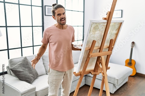 Young hispanic man with beard painting on canvas at home angry and mad screaming frustrated and furious, shouting with anger. rage and aggressive concept.