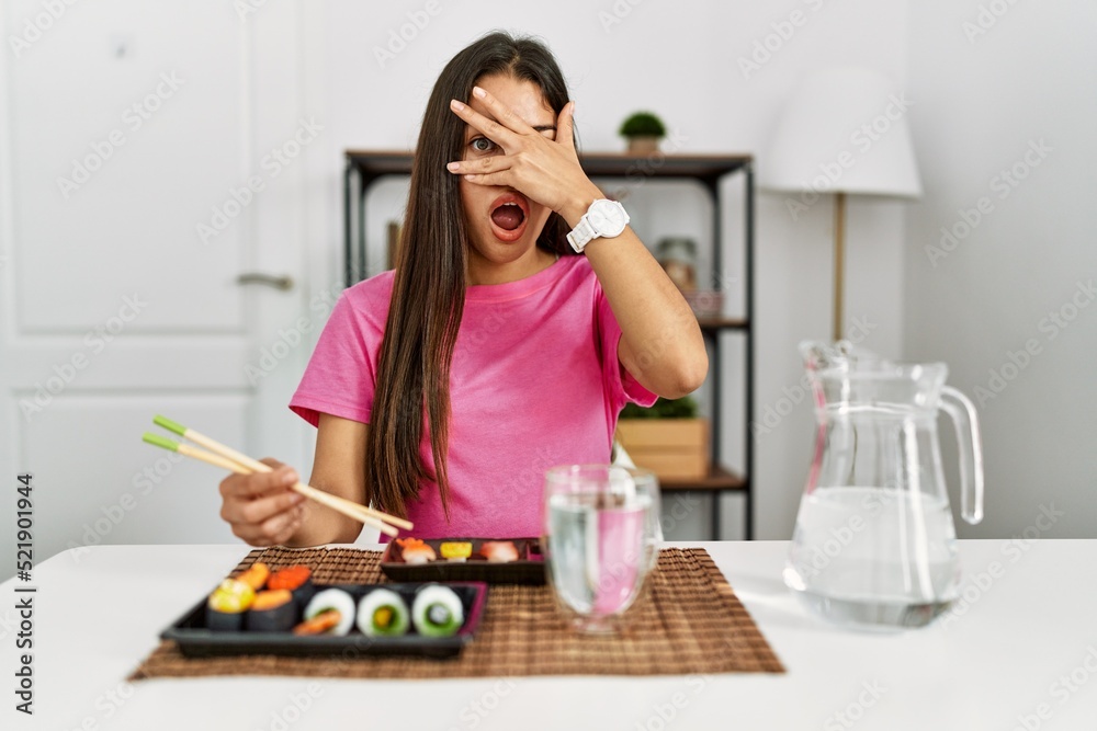 Young brunette woman eating sushi using chopsticks peeking in shock covering face and eyes with hand, looking through fingers with embarrassed expression.