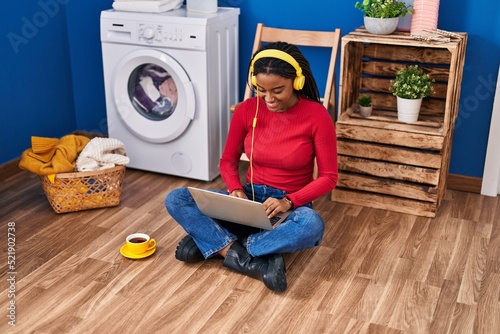 African american woman using laptop waiting for washing machine at laundry room