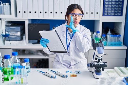Hispanic young woman working at scientist laboratory hand on mouth telling secret rumor  whispering malicious talk conversation