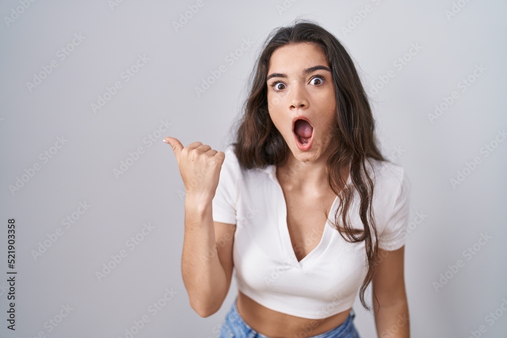 Young teenager girl standing over white background surprised pointing with hand finger to the side, open mouth amazed expression. Young teenager girl standing over white background surprised pointing with hand finger to the side, open mouth amazed expression.