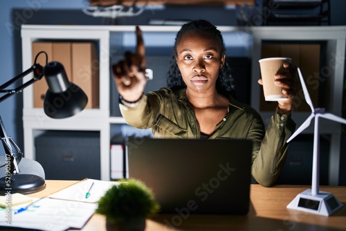 African woman working using computer laptop at night pointing with finger up and angry expression, showing no gesture