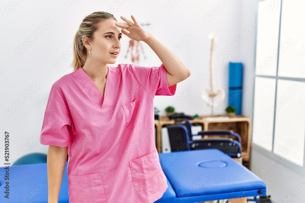 Young blonde woman working at pain recovery clinic very happy and smiling looking far away with hand over head. searching concept. Young blonde woman working at pain recovery clinic very happy and smiling looking far away with hand over head. searching concept.