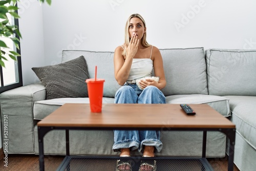 Young blonde woman eating popcorn sitting on the sofa covering mouth with hand, shocked and afraid for mistake. surprised expression