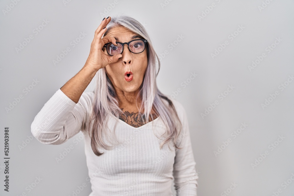 Middle age woman with grey hair standing over white background doing ok gesture shocked with surprised face, eye looking through fingers. unbelieving expression. Middle age woman with grey hair standing over white background doing ok gesture shocked with surprised face, eye looking through fingers. unbelieving expression.