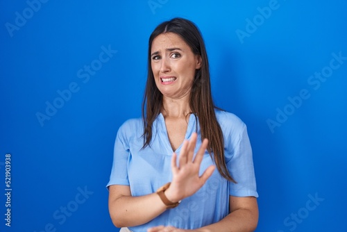 Young brunette woman standing over blue background disgusted expression, displeased and fearful doing disgust face because aversion reaction.