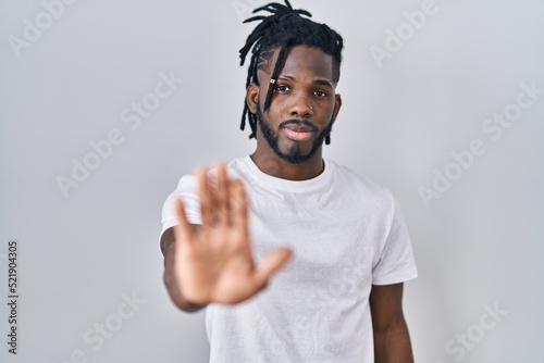 African man with dreadlocks wearing casual t shirt over white background doing stop sing with palm of the hand. warning expression with negative and serious gesture on the face.