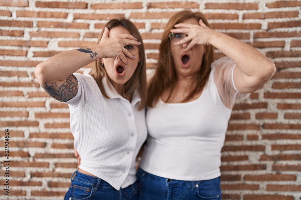 Hispanic mother and daughter wearing casual white t shirt peeking in shock covering face and eyes with hand, looking through fingers with embarrassed expression.