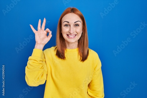 Young woman standing over blue background smiling positive doing ok sign with hand and fingers. successful expression. © Krakenimages.com