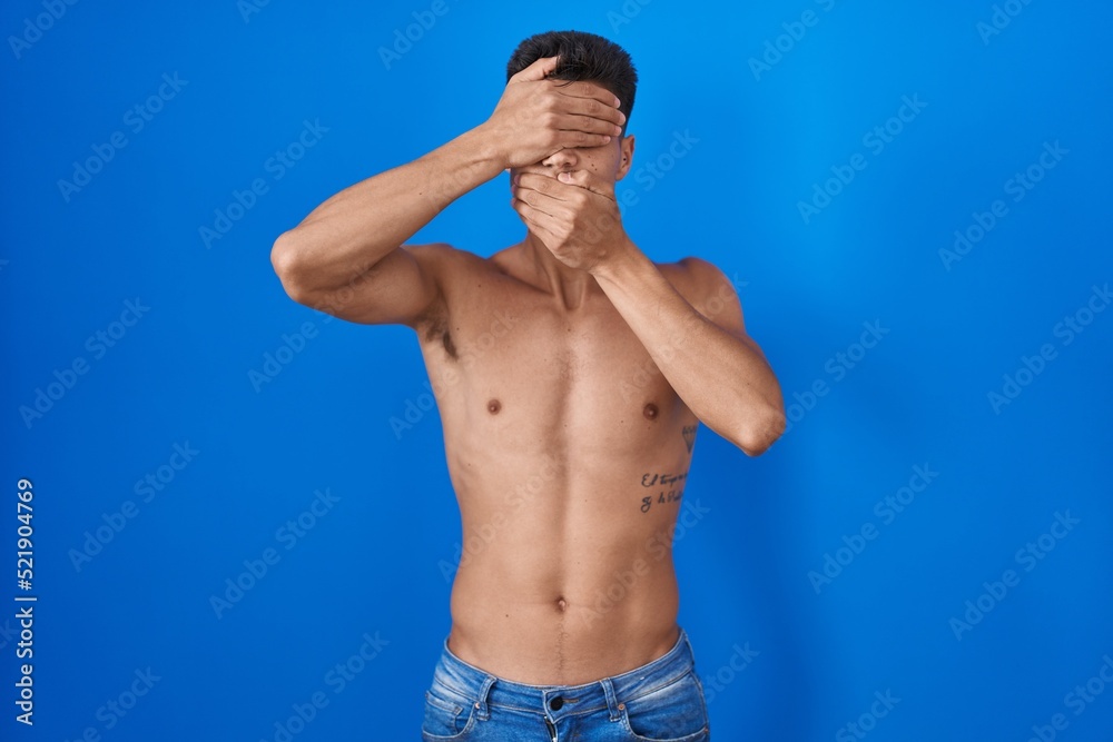 Young hispanic man standing shirtless over blue background covering eyes and mouth with hands, surprised and shocked. hiding emotion