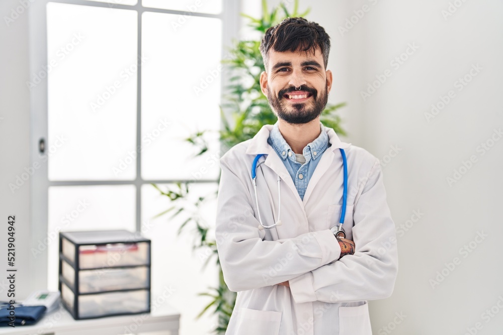 Young hispanic man wearing doctor uniform standing with arms crossed gesture at clinic