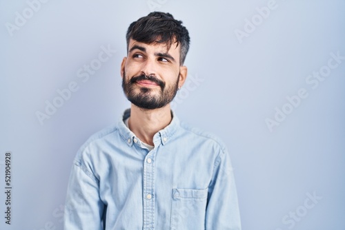 Young hispanic man with beard standing over blue background smiling looking to the side and staring away thinking. © Krakenimages.com