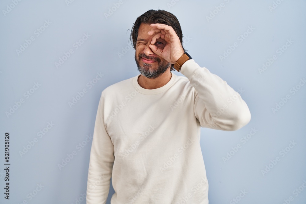 Handsome middle age man wearing casual sweater over blue background doing ok gesture with hand smiling, eye looking through fingers with happy face.