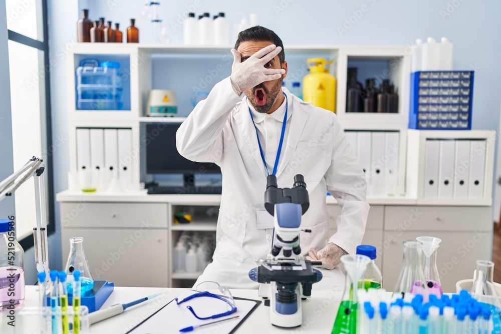 Young hispanic man with beard working at scientist laboratory peeking in shock covering face and eyes with hand, looking through fingers with embarrassed expression. Young hispanic man with beard working at scientist laboratory peeking in shock covering face and eyes with hand, looking through fingers with embarrassed expression.