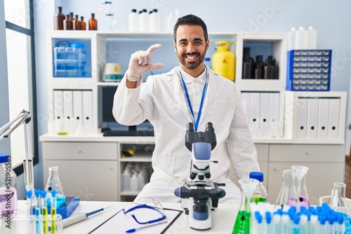 Young hispanic man with beard working at scientist laboratory smiling and confident gesturing with hand doing small size sign with fingers looking and the camera. measure concept.