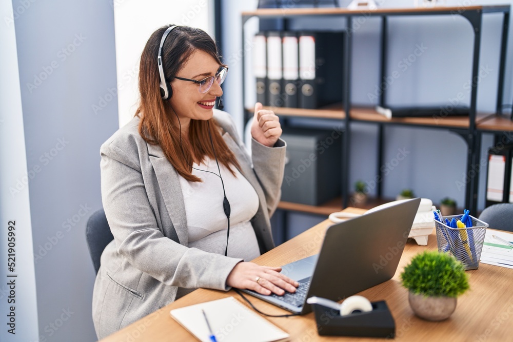 Pregnant woman working at the office wearing operator headset pointing thumb up to the side smiling happy with open mouth Pregnant woman working at the office wearing operator headset pointing thumb up to the side smiling happy with open mouth