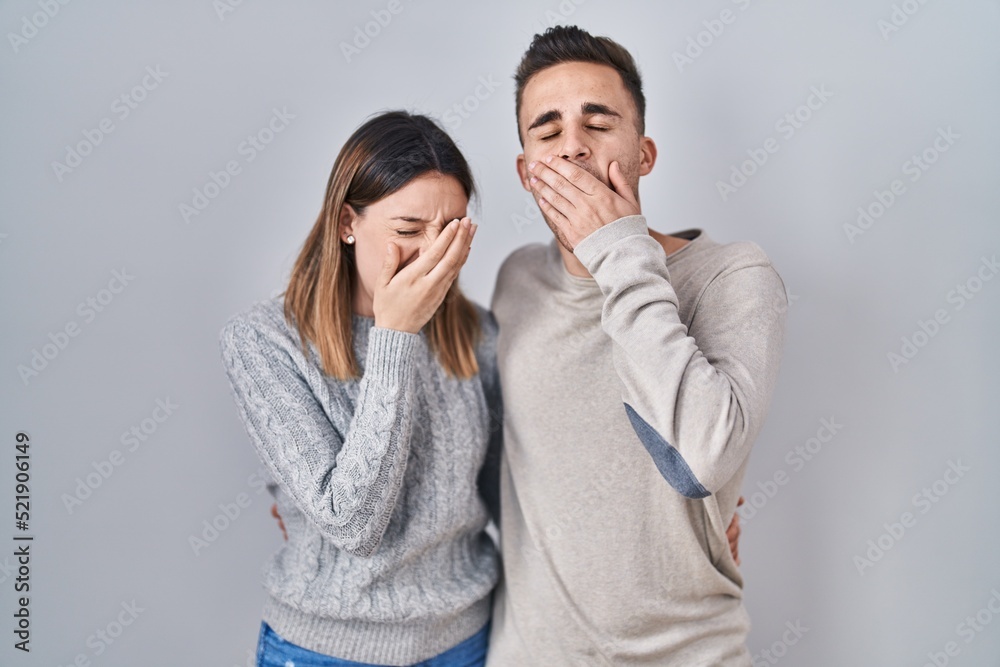 Young hispanic couple standing over white background bored yawning tired covering mouth with hand. restless and sleepiness.