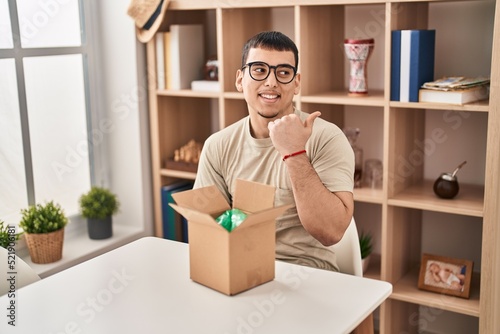 Young arab man with open gift in cardboard box pointing thumb up to the side smiling happy with open mouth