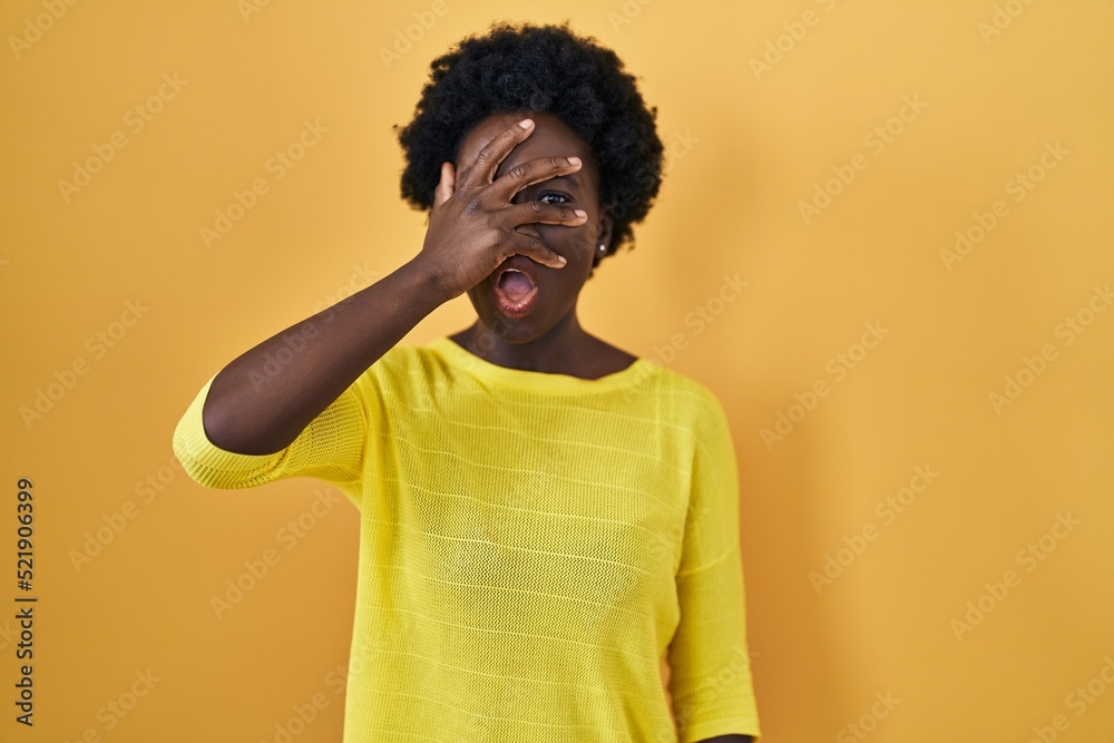 African young woman standing over yellow studio peeking in shock covering face and eyes with hand, looking through fingers with embarrassed expression.