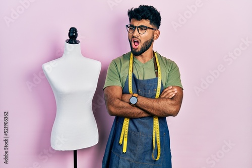 Arab man with beard dressmaker designer wearing atelier apron angry and mad screaming frustrated and furious, shouting with anger. rage and aggressive concept.