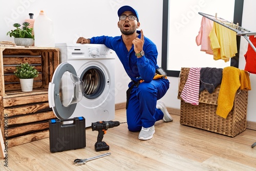Young indian technician working on washing machine amazed and surprised looking up and pointing with fingers and raised arms.