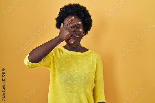 African young woman standing over yellow studio peeking in shock covering face and eyes with hand  looking through fingers with embarrassed expression.