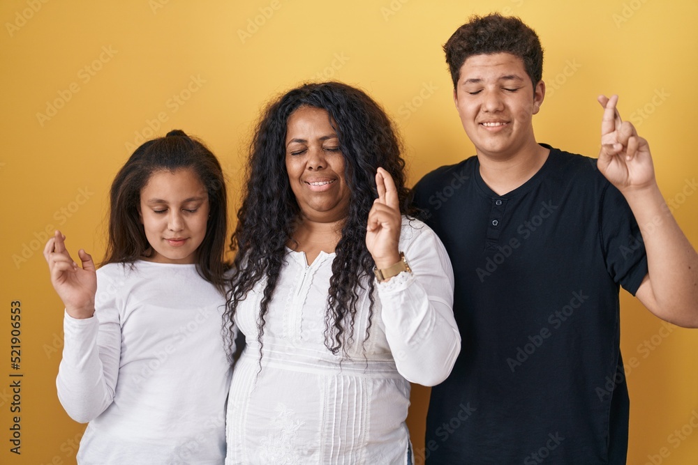 Family of mother, daughter and son standing over yellow background gesturing finger crossed smiling with hope and eyes closed. luck and superstitious concept. Family of mother, daughter and son standing over yellow background gesturing finger crossed smiling with hope and eyes closed. luck and superstitious concept.