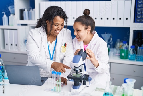 Mother and daughter scientists using microscope and laptop working at laboratory