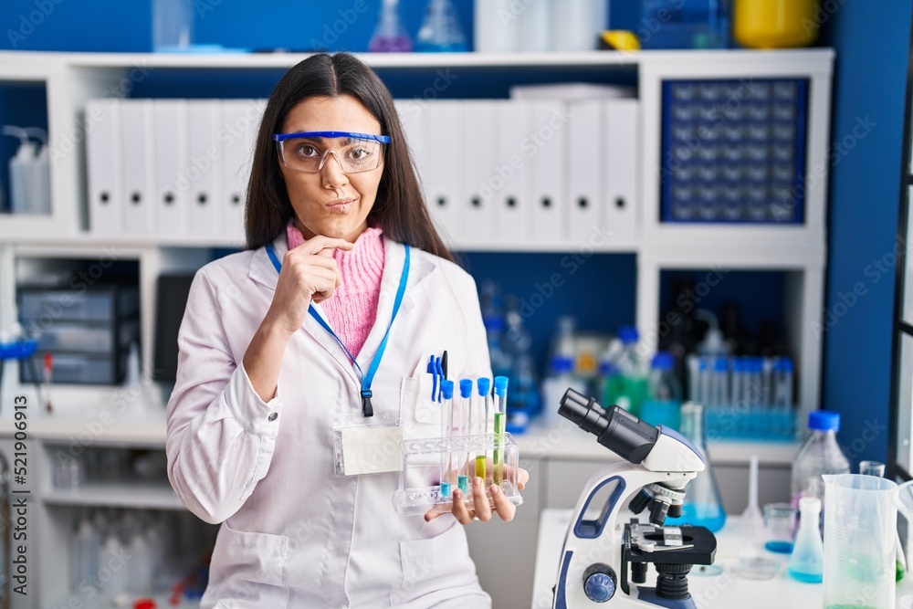 Young brunette woman working at scientist laboratory serious face thinking about question with hand on chin, thoughtful about confusing idea