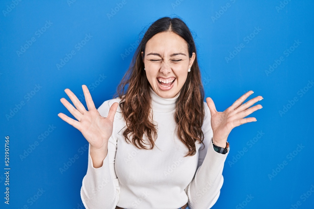 Fototapeta premium Young hispanic woman standing over blue background celebrating mad and crazy for success with arms raised and closed eyes screaming excited. winner concept