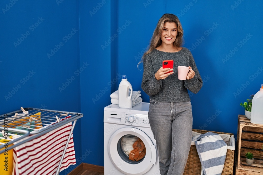 Young blonde woman using smartphone and drinking coffee waiting for washing machine at laundry room