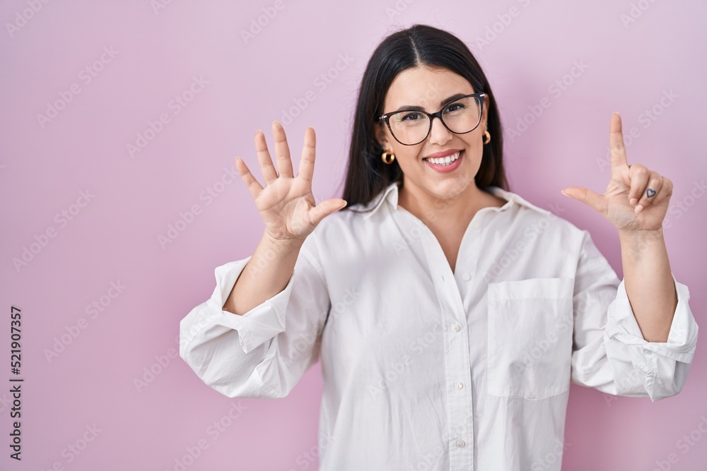 Young brunette woman standing over pink background showing and pointing up with fingers number seven while smiling confident and happy. Young brunette woman standing over pink background showing and pointing up with fingers number seven while smiling confident and happy.