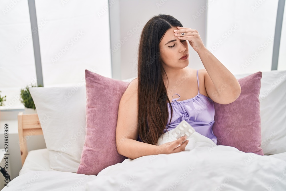 Young hispanic woman taking pills for headache sitting on bed at bedroom