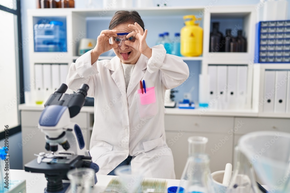 Fototapeta premium Hispanic girl with down syndrome working at scientist laboratory doing ok gesture like binoculars sticking tongue out, eyes looking through fingers. crazy expression.