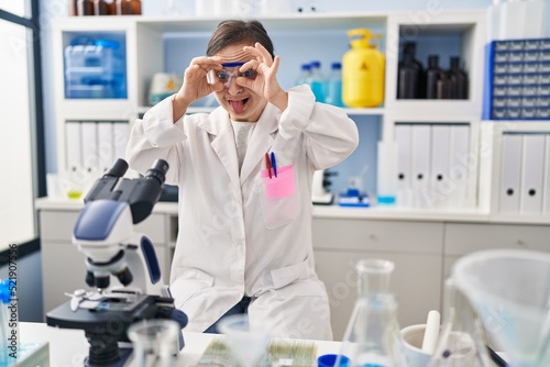 Hispanic girl with down syndrome working at scientist laboratory doing ok gesture like binoculars sticking tongue out, eyes looking through fingers. crazy expression.