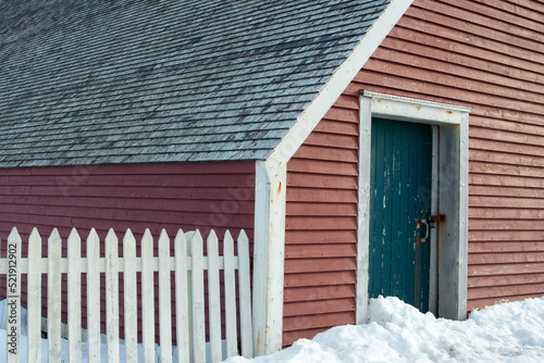 The exterior of a red wooden cape cod clapboard or batten board siding wall of a country style cottage with a white wooden picket fence in the foreground. The roof has wooden cedar shake shingles. photo