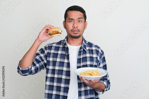 Adult Asian man holding a burger and french fries with serious face expression photo