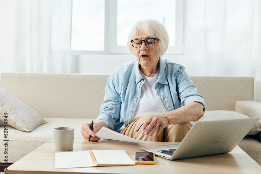 a pleasant elderly lady, passionate about work, sits in her apartment on a comfortable sofa in stylish clothes and uses a laptop and looks at her notebook for notes