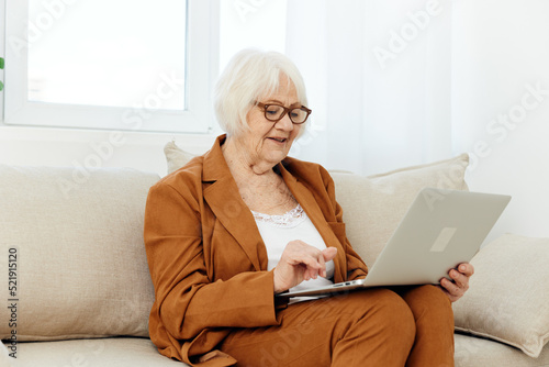 Mila is a joyful old lady sitting with a laptop on her lap on a beige sofa near the window and looking at the monitor with a pleasant smile on her face working after retirement