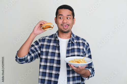 Asian man showing funny tempted expression when looking to burger that he hold photo