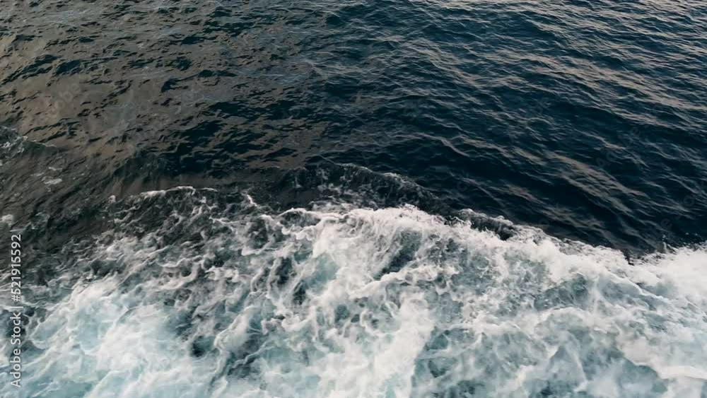 Large boat glides through the sea creating a wake of hypnotic waves. The sunlight glistens on the surface of the water. Water flows into waves against the navy blue sea water in slow motion. Large boat glides through the sea creating a wake of hypnotic waves. The sunlight glistens on the surface of the water. Water flows into waves against the navy blue sea water in slow motion.