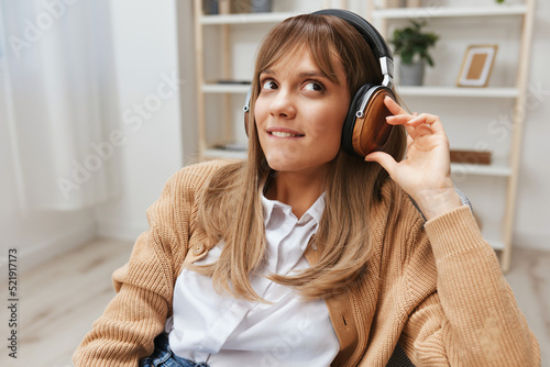 Dreaming young blonde student lady freelancer in warm sweater in headphones listen fav songs looks aside sitting in armchair at home interior. Music time Relaxing Cool playlist Concept. Copy space photo