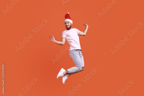 Portrait of funny bearded man in santa hat jumping in the air and looking at camera with positive smile, spreading hands aside, wearing pink T-shirt. Indoor studio shot isolated on orange background.