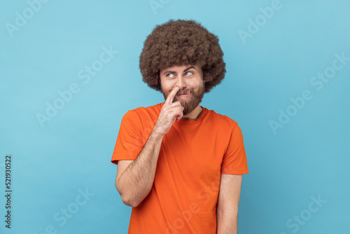 Portrait of childish man with Afro hairstyle wearing orange T-shirt picking nose and sticking out tongue with comical stupid expression. Indoor studio shot isolated on blue background.