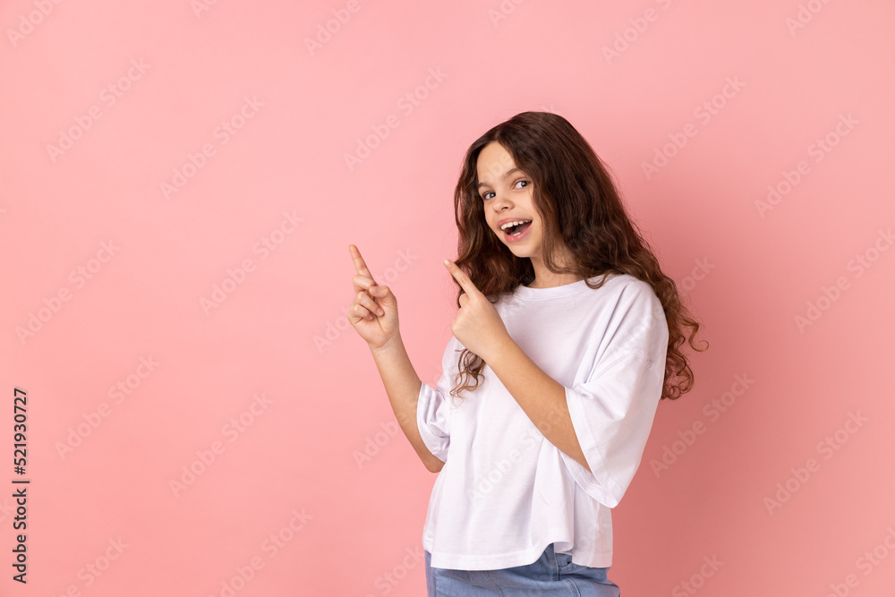 Portrait of delighted little girl wearing white T-shirt pointing fingers aside to advertising area, copy space for advertisement. Indoor studio shot isolated on pink background.