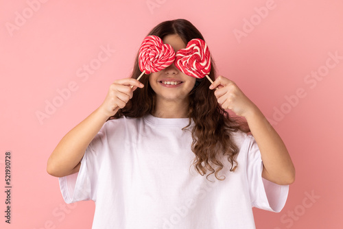 Portrait of little girl wearing white T-shirt covering her eyes with heart shape candies and smiling happily, posing with pout lips. Indoor studio shot isolated on pink background.